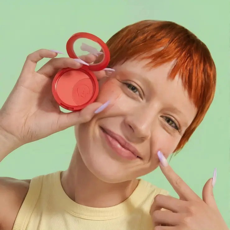 Playful lifestyle cosmetic ad featuring a smiling AI virtual model with short red hair holding an open coral blush compact against a light green background
